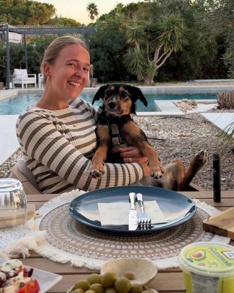 Eileen lacht in die Kamera und hält Klette, einen kleinen schwarz-braunen Hund, auf dem Schoß. Im Hintergrund ein Garten mit Pool.